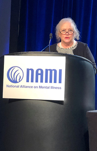 Image of Judge Joyce Campbell standing at a podium in front of a blue curtain with a podium sign that says NAMI