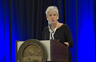 Image of Chief Justice Maureen O'Connor standing at a podium, with a blue curtain in the background