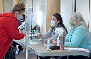 Image of two women sitting at a table, facing another women behind a plexiglass barrier.