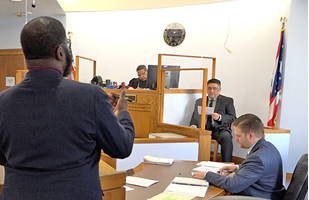 A courtroom with a Black male judge on a bench, an Asian male man in the witness box, and Black male attorney standing in front of them.