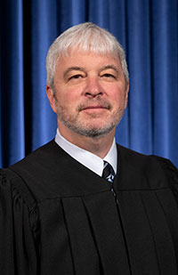 Image is a headshot photo of Justice Michael Donnelly in his black judicial robe with a blue pleated curtain in the background