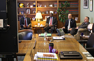 Image of four men dressed in suits sitting in a conference room facing a large monitor