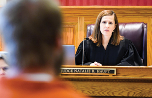 Image of an inmate wearing an orange jumpsuit standing before a female judge in a courtroom