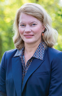 Image is a head shot of a blond woman with greenery in the background