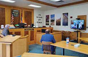 Two men sitting in wooden chairs watching a video monitor in a courtroom