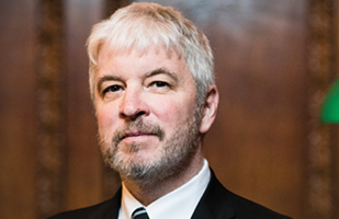 Image is a head shot of Justice Donnelly in a dark suit jacket, white dress shirt, and dark tie