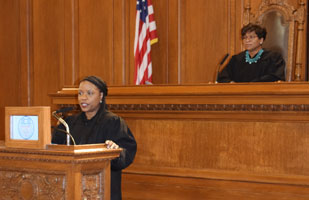 Image of a woman wearing a black judicial robe speaking from behind a podium. Behind her sits another woman wearing a black judicial robe