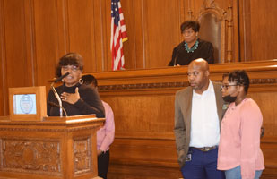 Image of a woman speaking from behind a podium. A man and woman are standing next to her and another woman wearing a black judicial robe is sitting behind her