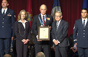 Image of a woman in a dark pant suit standing next to a man wearing a dark suit, adorned with military bars. He is wearing a medalion around his neck and holding a plaque. He is standing next to Ohio Goveror Mike DeWine. Two men wearing military dress blues stand at attention on either side of these three people
