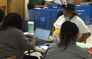 Image of two women sitting at a table behind plexiglass partitions assisting a man