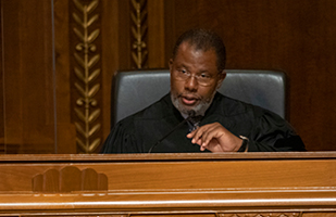 Image of a male judge wearing a black judicial robe sitting at a wooden judicial bench