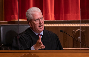 White man with grey hair and glasses wearing a black judicial robe sitting behind a bench in front of microphone.