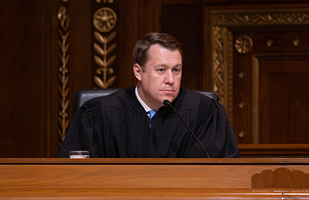 Image of a male judge wearing a black judicial robe sitting at a wooden bench in the courtroom of the Thomas J. Moyer Ohio Judicial Center.