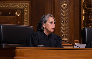 Image of a female judge wearing a black judicial robe sitting at a wooden bench in the courtroom of the Thomas J. Moyer Ohio Judicial Center.