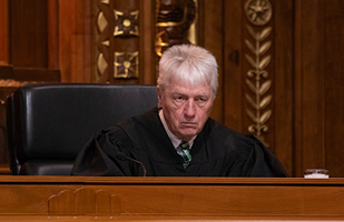 Image of a male judge wearing a black judicial robe sitting at a wooden bench in the courtroom of the Thomas J. Moyer Ohio Judicial Center.