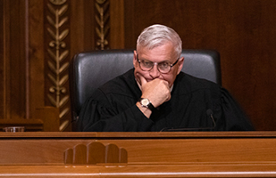 Image of a male judge wearing a black judicial robe sitting in a black, leather chair at a wooden bench in the courtroom of the Thomas J. Moyer Ohio Judicial Center.