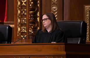 Image of a female judge wearing black framed glasses and a black judicial robe seated at a wooden courtroom bench.