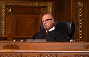 Image of a male judge wearing a black judicial robe sitting at a wooden courtroom bench.