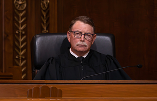 Image of a male judge wearing a black judicial robe seated at a wooden bench in the courtroom of the Thomas J. Moyer Ohio Judicial Center.