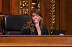 Image of a female judge wearing a black judicial robe sitting at an intricately carved wooden bench listening to oral arguments in the courtroom of the Thomas J. Moyer Ohio Judicial Center.