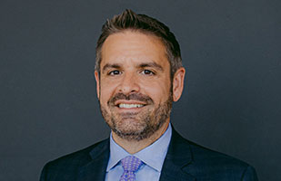 White man with a beard wearing a navy suit, light blue shirt, and purple tie.