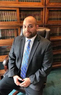 Image of a man wearing a grey suit sitting in a leather office chair in front of a wall of law books