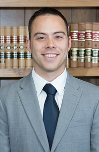 Image of a man with short, dark hair, wearing a grey suit and blue tie standing in front of a bookcase of law books