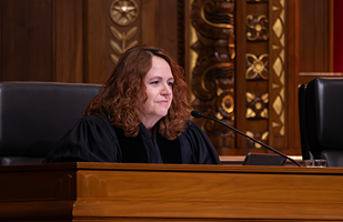 Image of a female judge with long, red, curly hair, wearing a black judicial robe seated at a wooden courtroom bench.