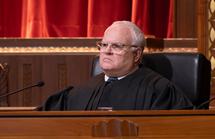 Image of white male with glasses in a judicial robe behind a bench.