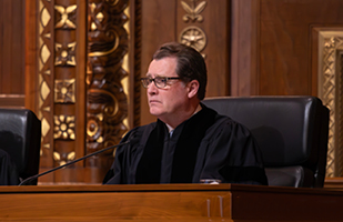 Image of a male judge wearing a black judicial robe sitting at a wooden bench in the courtroom of the Thomas J. Moyer Ohio Judicial Center.