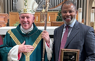 A white Catholic bishop standing next to a Black man in a suit and tie with an award.