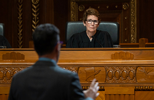 A woman with short, brown hair and glasses wearing a black judicial robe sits at a wooden bench listening to a man wearing a suit standing before her in the courtoom of the Thomas J. Moyer Ohio Judicial Center.