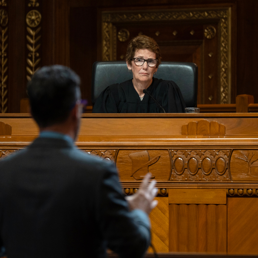 A woman with short, brown hair and glasses wearing a black judicial robe sits at a wooden bench listening to a man wearing a suit standing before her in the courtoom of the Thomas J. Moyer Ohio Judicial Center.