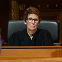 A woman with short, brown hair and glasses wearing a black judicial robe sits at a wooden bench listening to a man wearing a suit standing before her in the courtoom of the Thomas J. Moyer Ohio Judicial Center.