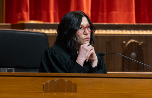A woman with long, dark hair, wearing glasses and a black judicial robe seated behind a wooden bench in the courtroom of the Thomas J. Moyer Ohio Judicial Center.