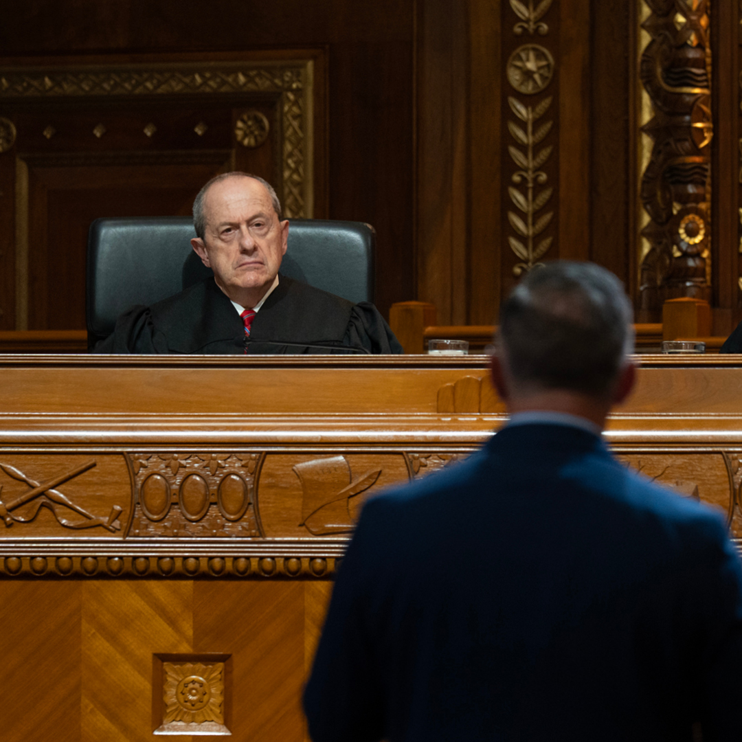 A man wearing a black judicial robe is seated at a wooden bench looking at a man wearing a blue suit standing in front of the bench in the courtroom of the Thomas J. Moyer Ohio Judicial Center.