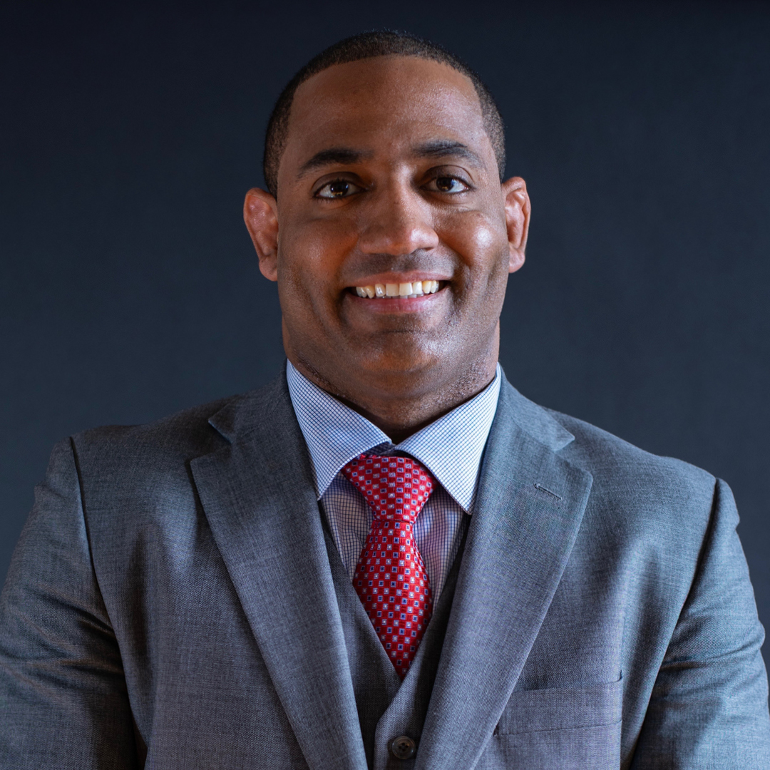 A man wearing a grey three-piece suit, white dress shirt, and red tie.