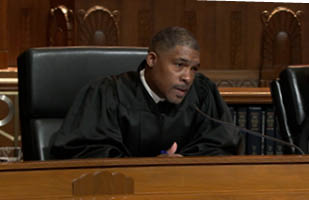 A man sitting at a bench speaking into a microphone wearing judicial robes.