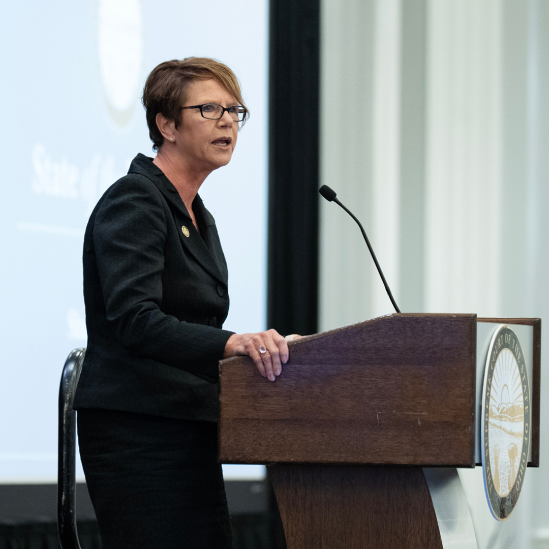A woman wearing a dark suit speaks from a wooden podium.