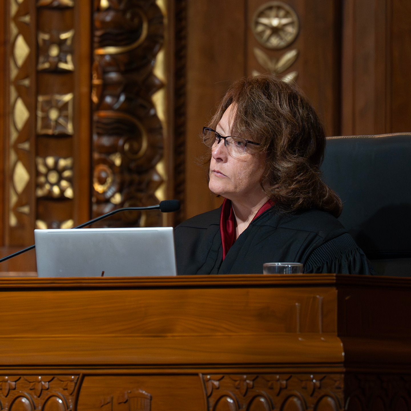 A female judge in a black robe sits at a wooden bench with ornate carvings, focused and looking to her right, with a laptop.