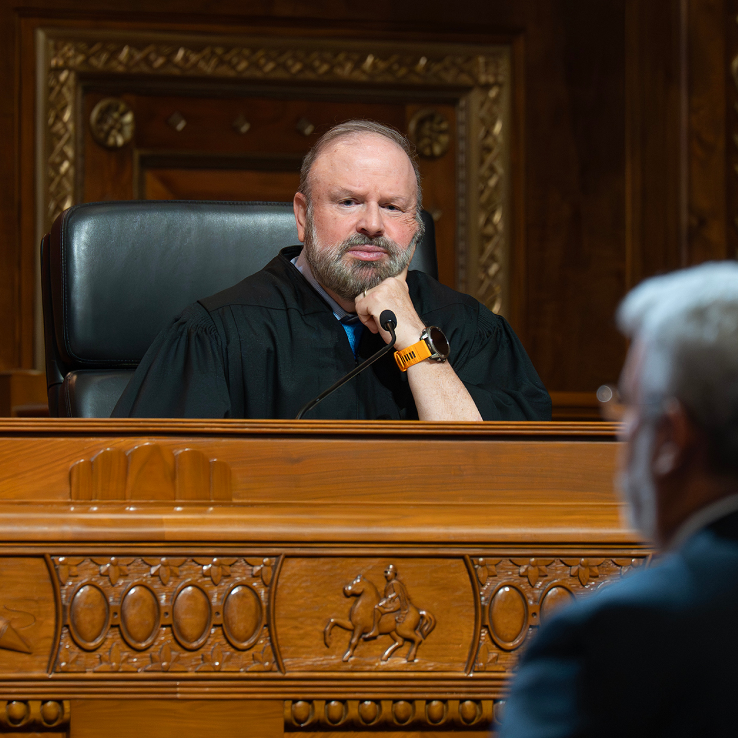 A judge in a black robe sitting at a wooden bench with his fist on his chin