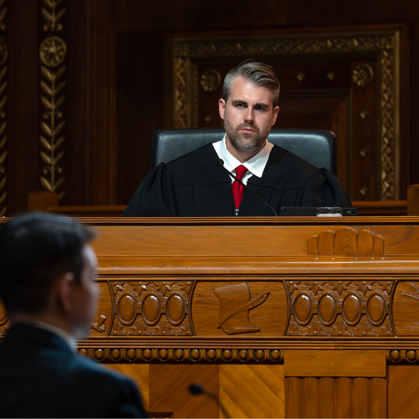 A judge in a black robe, sits behind an ornate wooden bench in a courtroom, facing a blurred person in the foreground.