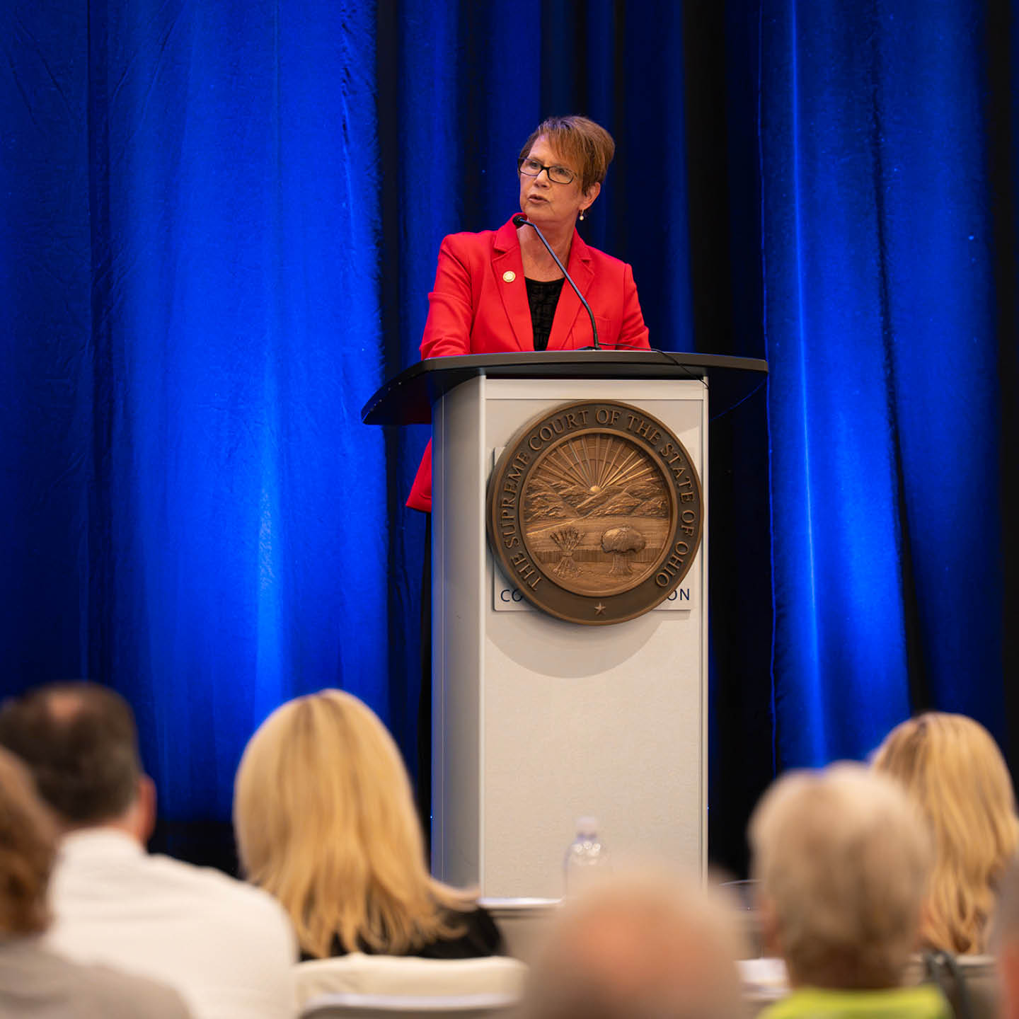 A woman with short brown hair and glasses, wearing a bright red blazer over a black top, speaking at a podium.
