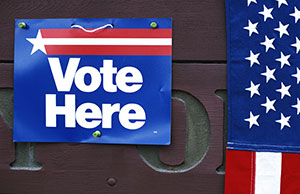 Image shows a "Vote Here" sticker and an American flag on a ballot box