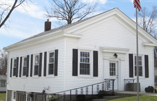 Image is of a white building with black shutters, with a black railing and am American flag by the front door