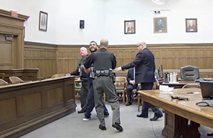 Image of a man wearing jail scrubs being restrained by police officers and court bailiffs in a courtroom.