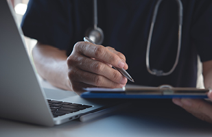 Image of a hand holding a pen and clipboard near a laptop. The person has a stethoscope around their neck.