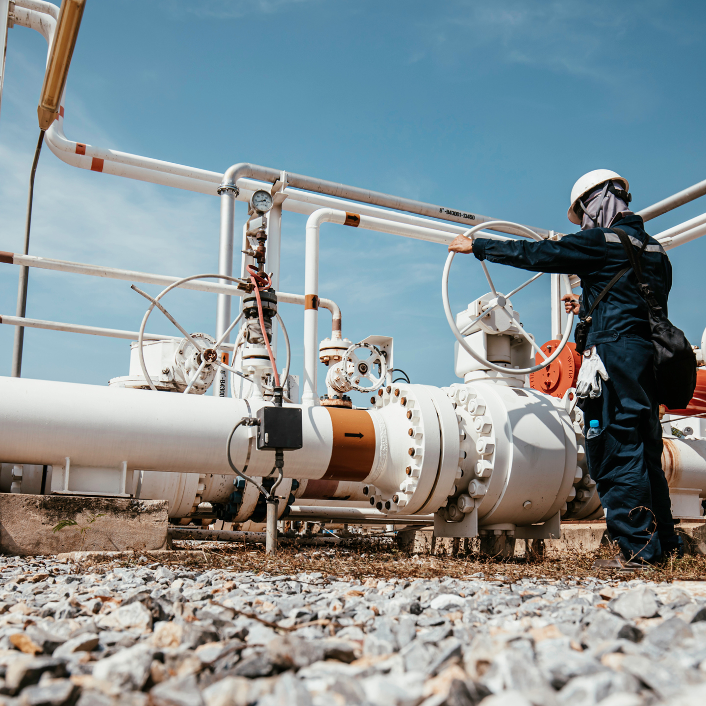 A worker wearing a blue jumpsuit and white hard hat working on a gas line.