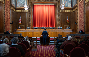 A man wearing a suit stands at a wooden podium facing seven men and women wearing black judicial robes seated at a wooden bench in the courtroom of the Thomas J. Moyer Ohio Judicial Center.