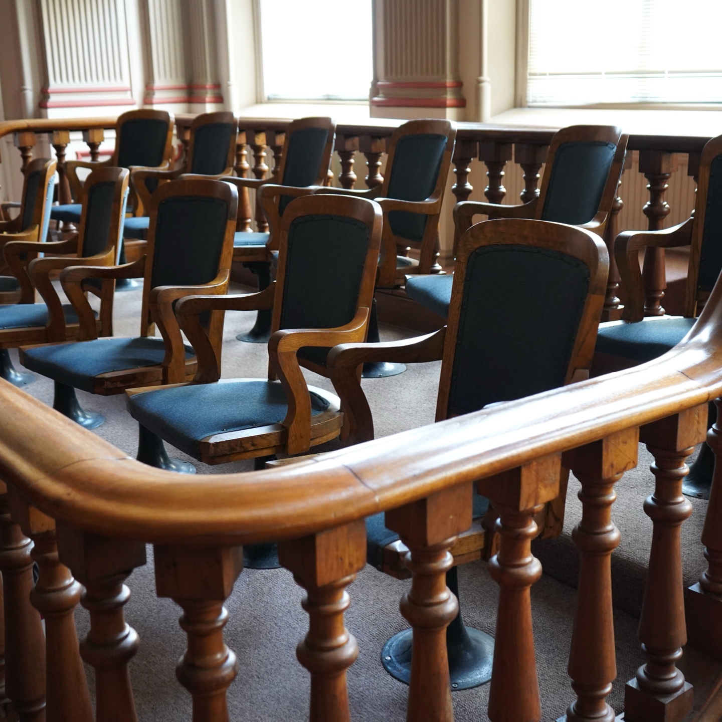 An angled, medium shot captures a jury box inside a courtroom. The area features rows of dark wooden, swivel chairs with dark blue upholstery. A low, curved wooden balustrade bounds the space. Windows fill the background.